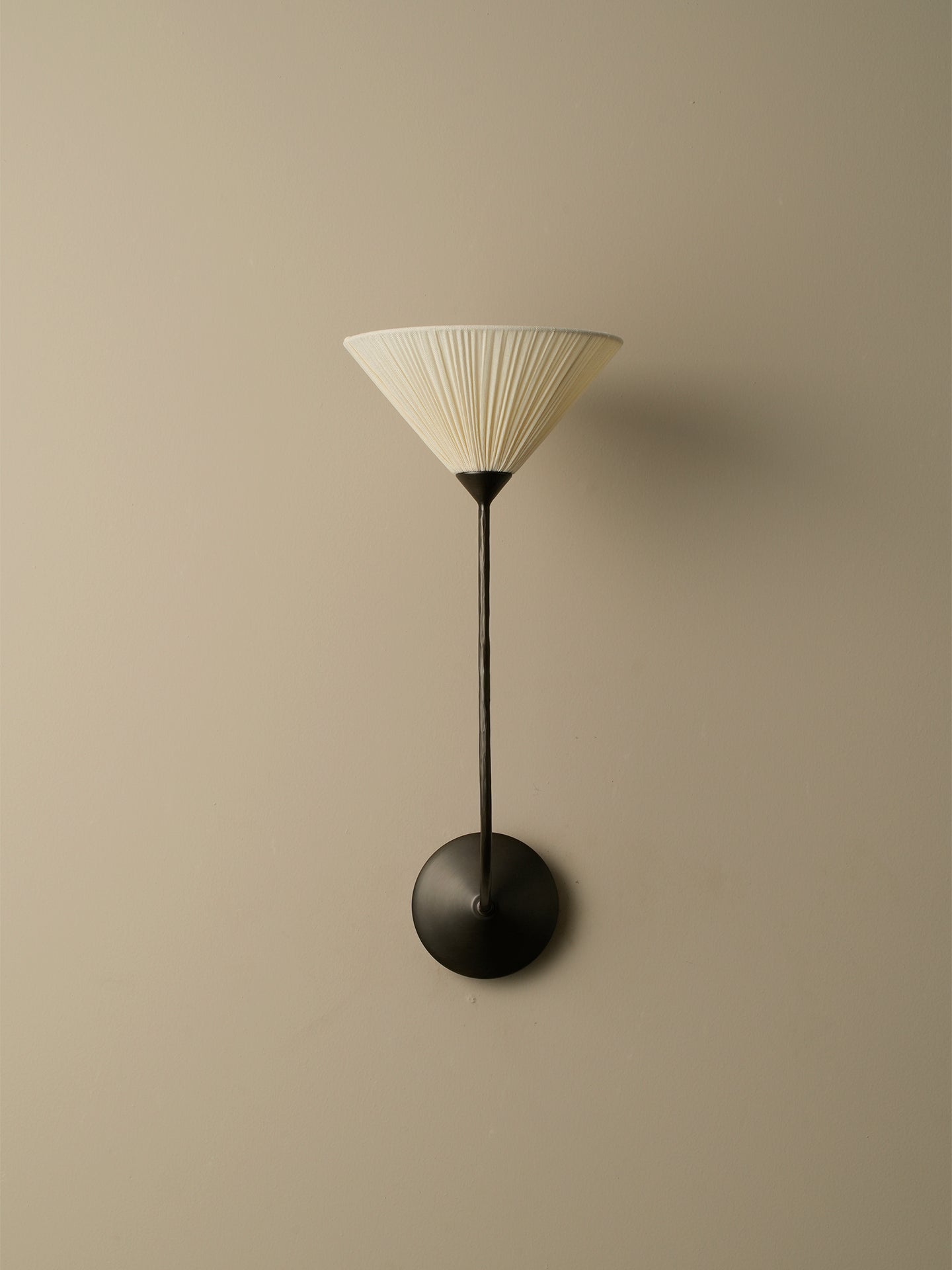A modern wall-mounted sconce with a pleated off-white conical shade, a slim dark metal stem, and a round dark metal base, shown against a plain beige wall.