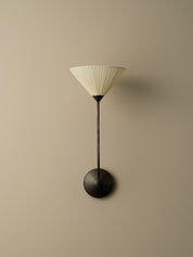 A modern wall-mounted sconce with a pleated off-white conical shade, a slim dark metal stem, and a round dark metal base, shown against a plain beige wall.