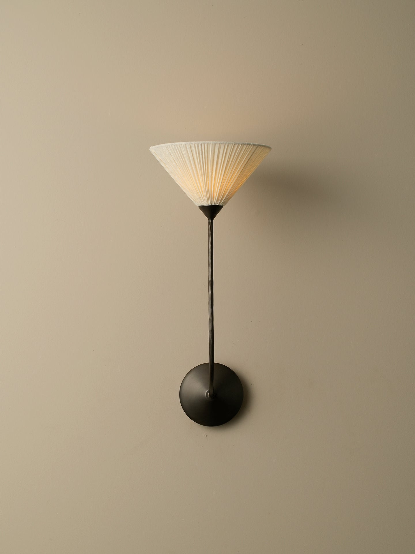 A modern wall sconce with a pleated white shade, a slim black stem, and a round black wall mount, shown against a plain beige wall.