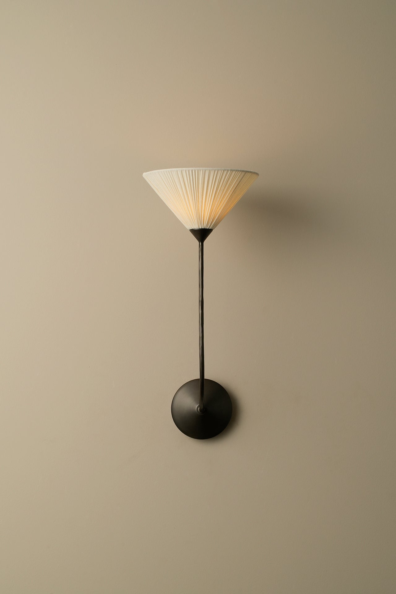 A modern wall-mounted sconce with a pleated white shade, a slim black stem, and a round black base, shown against a plain beige wall.