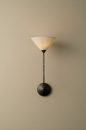 A modern wall-mounted sconce with a pleated white shade, a slim black stem, and a round black base, shown against a plain beige wall.