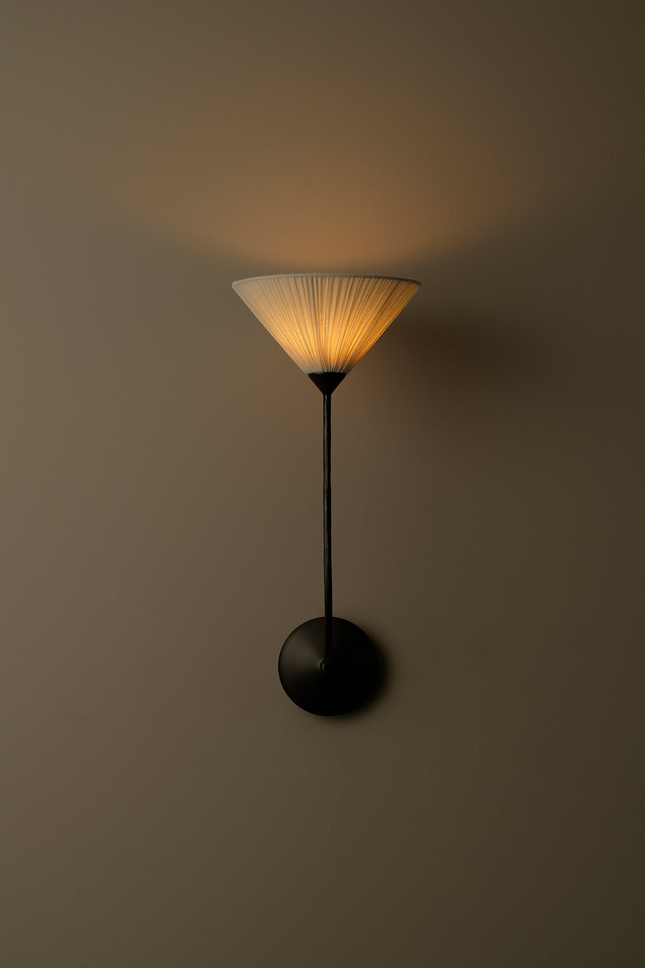 A modern wall-mounted sconce with a black metal stand and circular base, featuring a pleated white shade that glows with warm yellow light against a dark taupe wall.