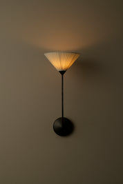 A modern wall-mounted sconce with a black metal stand and circular base, featuring a pleated white shade that glows with warm yellow light against a dark taupe wall.