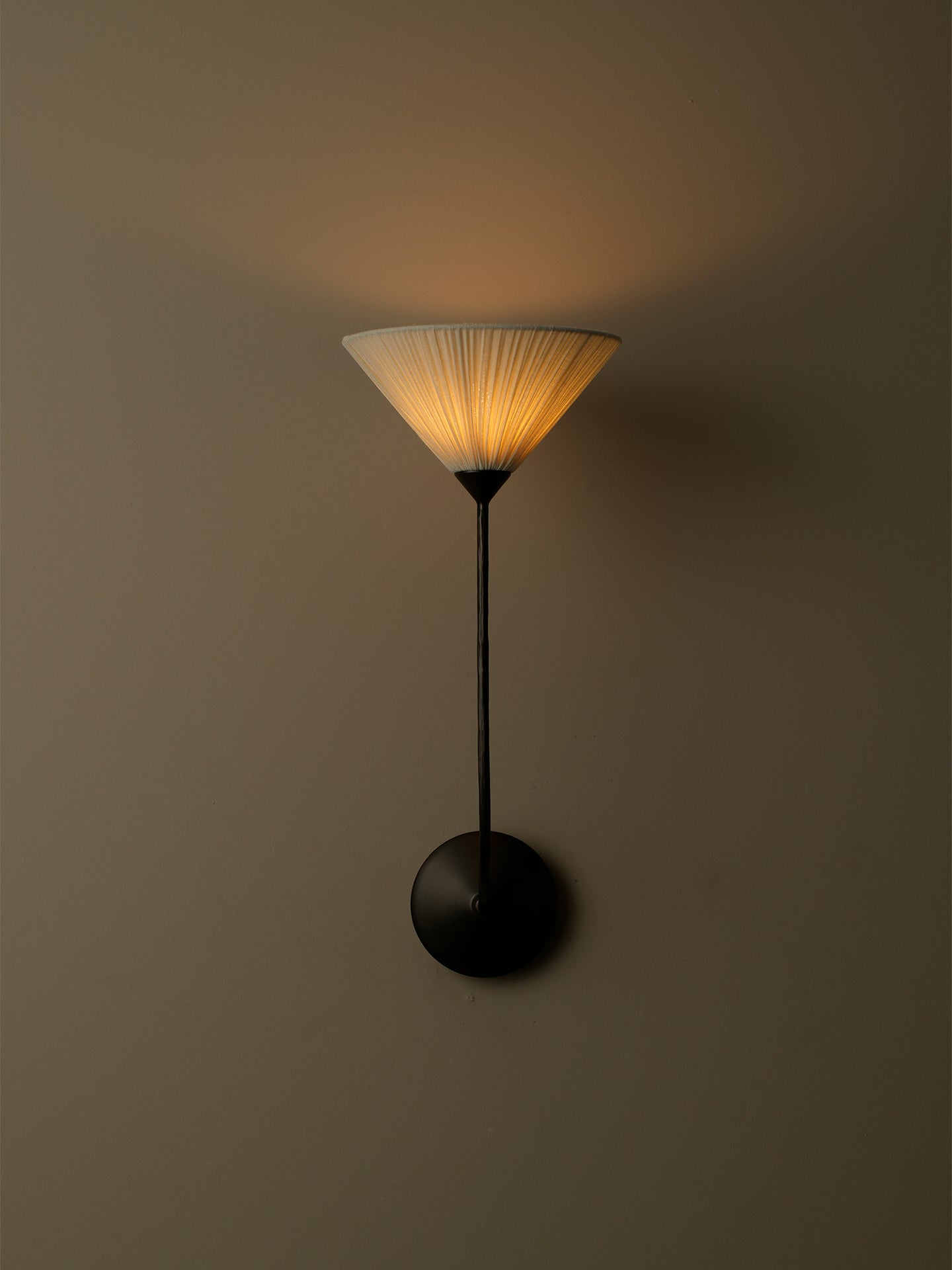 A modern wall sconce with a black metal stand and circular wall mount, featuring a pleated conical shade that glows warm yellow when lit, set against a dark neutral wall.