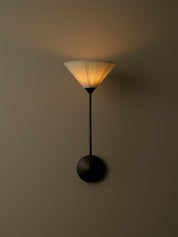 A modern wall sconce with a black metal stand and circular wall mount, featuring a pleated conical shade that glows warm yellow when lit, set against a dark neutral wall.