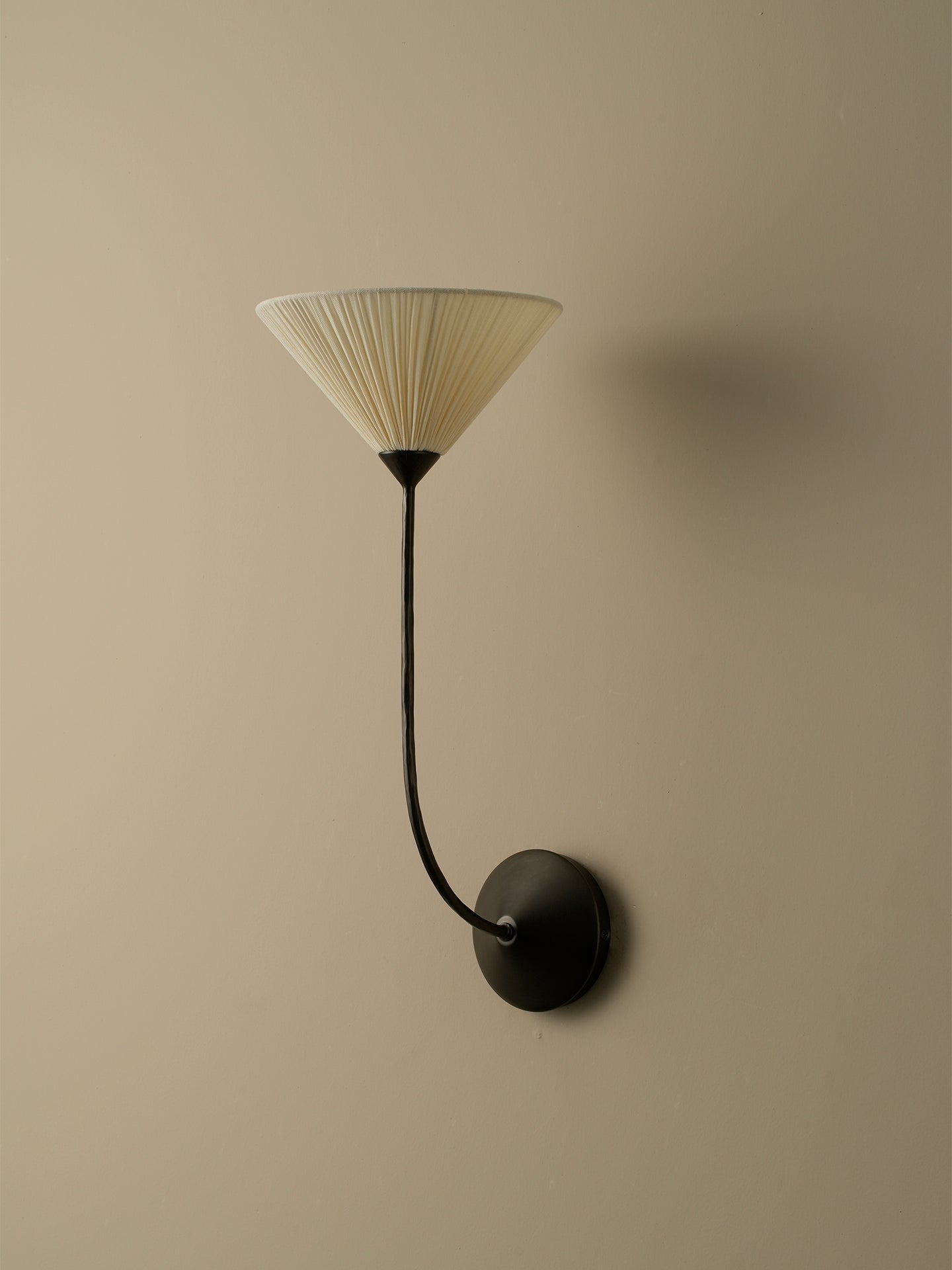 A modern wall sconce with a pleated cream shade and a curved black metal arm, mounted on a beige wall.