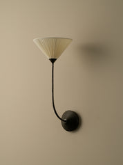 A modern wall sconce with a pleated cream shade and a curved black metal arm, mounted on a beige wall.