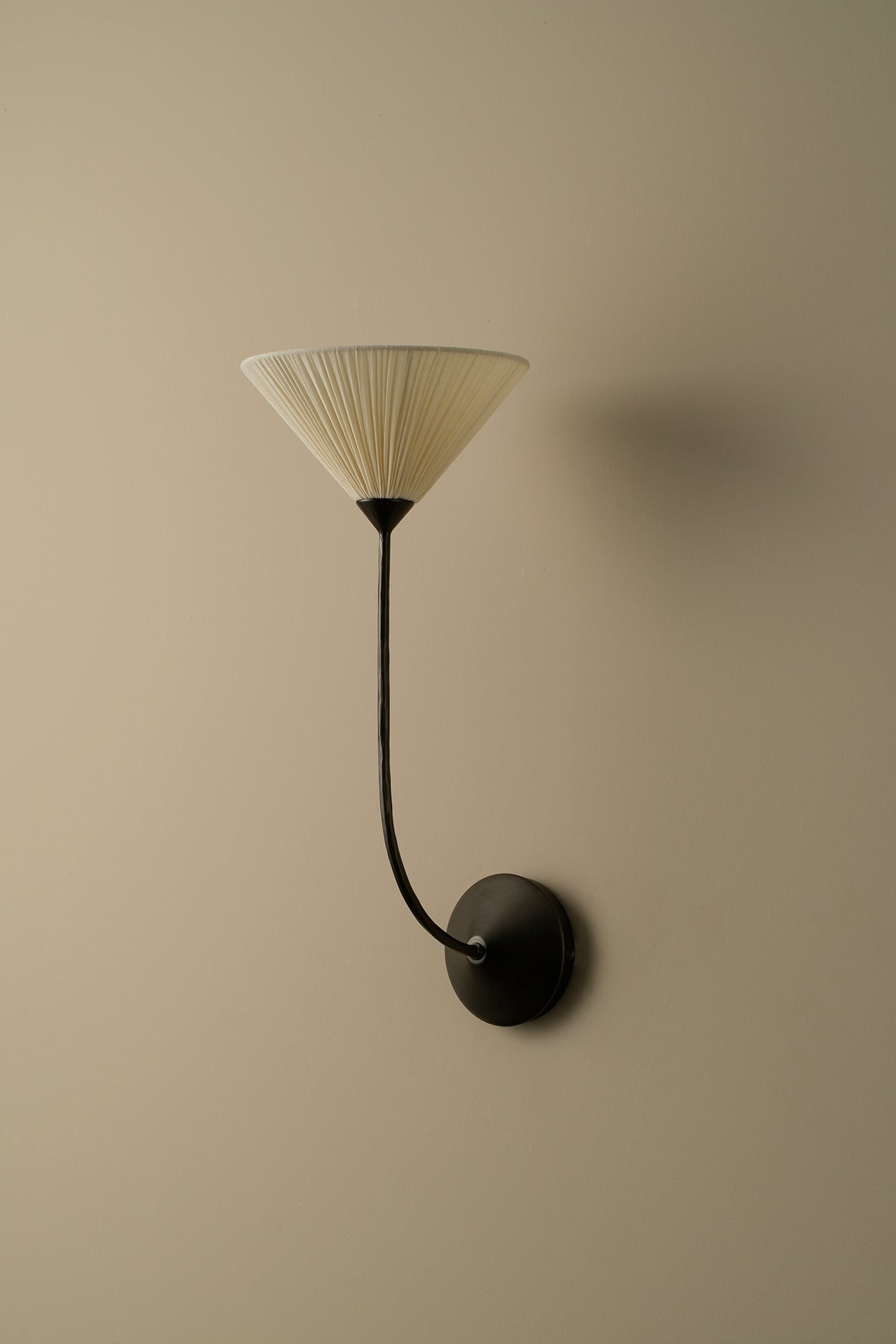 A modern wall-mounted sconce with a black curved arm and circular wall plate, featuring a pleated conical shade in warm off-white, mounted on a solid beige wall.