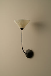 A modern wall-mounted sconce with a black curved arm and circular wall plate, featuring a pleated conical shade in warm off-white, mounted on a solid beige wall.