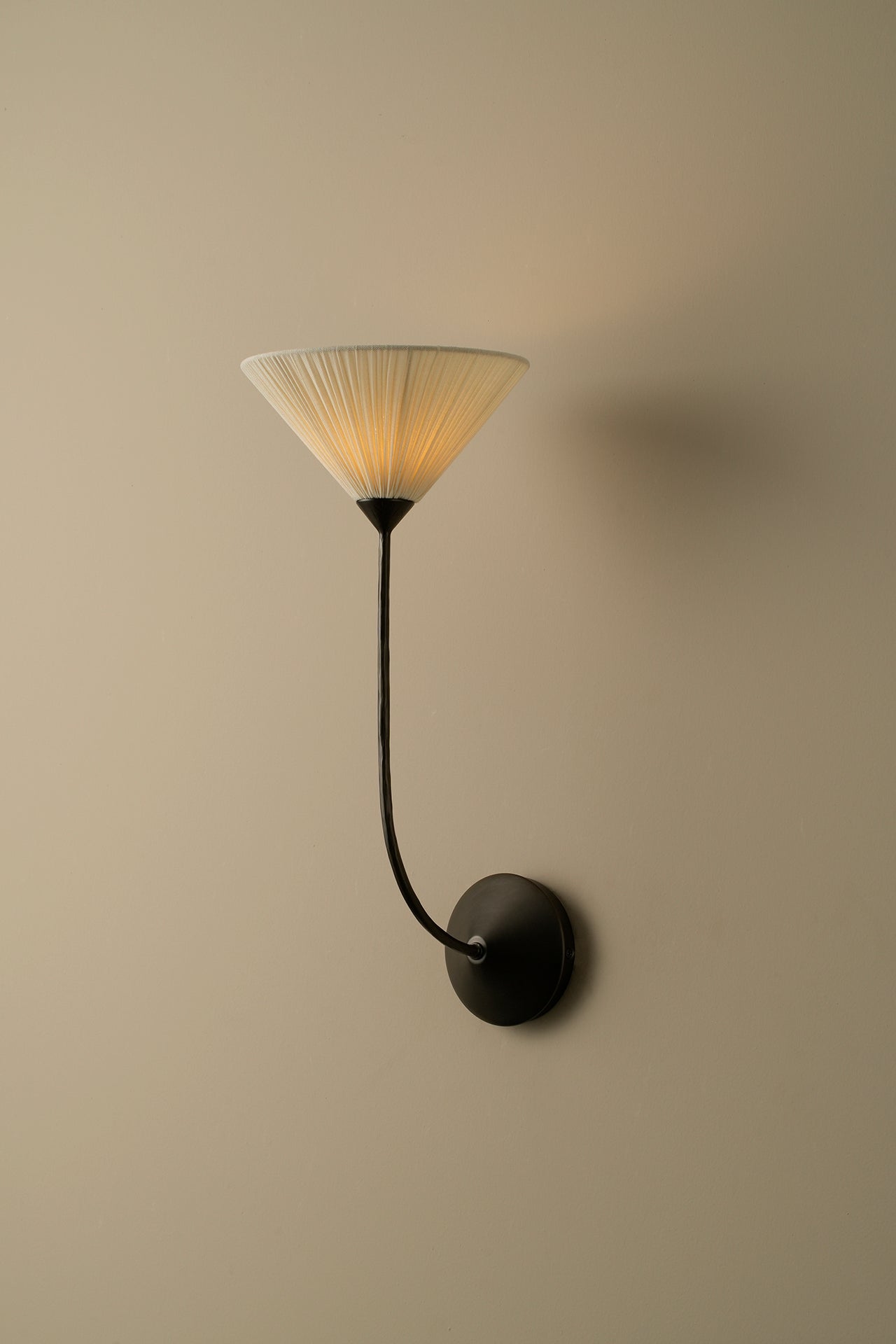 A modern wall sconce with a black curved arm and circular wall mount, featuring a pleated translucent shade that glows warm yellow when lit, set against a plain beige wall.
