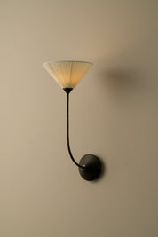 A modern wall sconce with a black curved arm and circular wall mount, featuring a pleated translucent shade that glows warm yellow when lit, set against a plain beige wall.