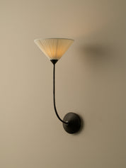 A modern wall sconce with a black curved arm and circular wall mount, featuring a pleated conical shade that glows with warm light against a beige wall.