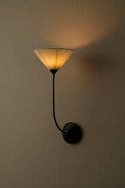 A modern wall sconce with a black metal curved arm and a pleated conical shade glowing warm yellow, mounted on a plain beige wall.