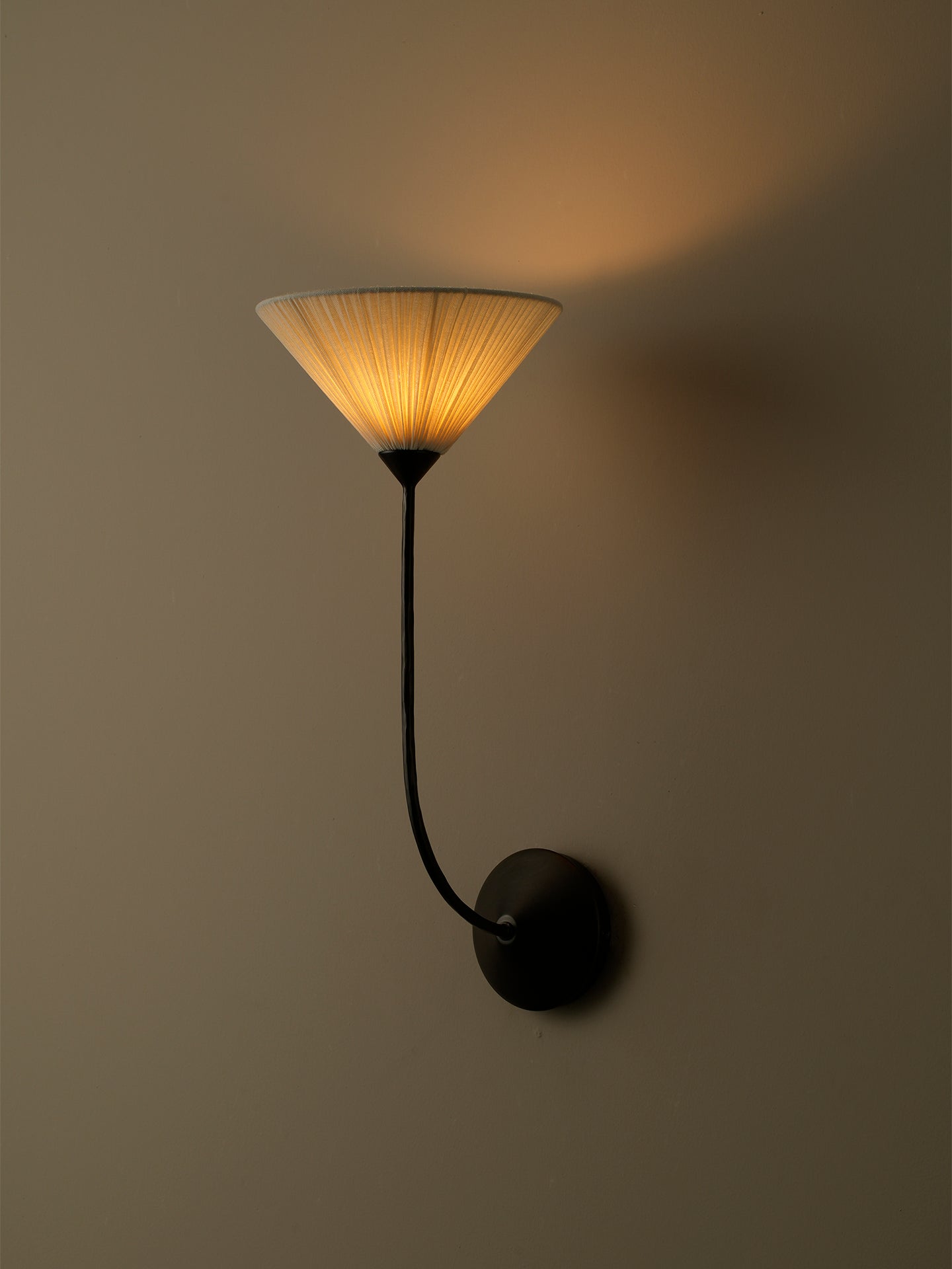 A modern wall sconce with a black metal curved arm and a pleated conical shade glowing with warm yellow light, mounted on a plain beige wall.
