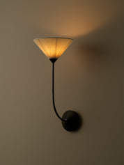 A modern wall sconce with a black metal curved arm and a pleated conical shade glowing with warm yellow light, mounted on a plain beige wall.