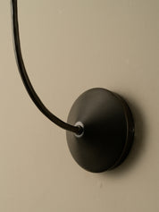 A close-up of a matte black wall-mounted sconce with a curved gooseneck arm, attached to a beige wall.