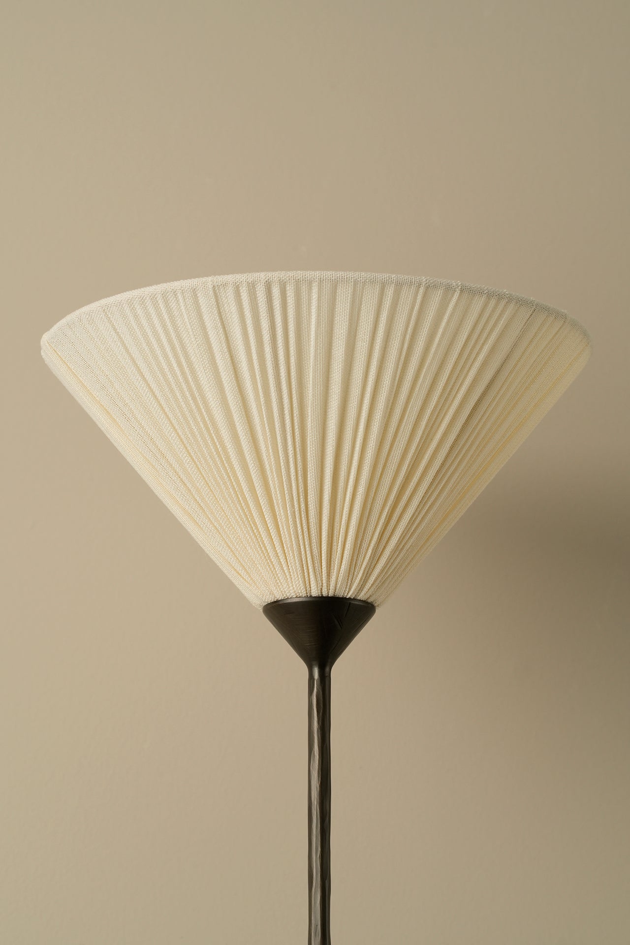 A pleated conical lampshade in warm off-white or cream, attached to a dark metal stand, set against a solid beige background.