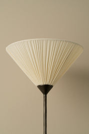 A pleated conical lampshade in warm off-white or cream, attached to a dark metal stand, set against a solid beige background.