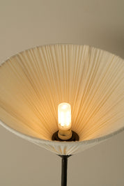 A close-up of a pleated fabric lampshade in a warm cream or off-white color, glowing with a single exposed filament light bulb inside. The lampshade is attached to a dark metal stand, set against a plain, warm beige background.