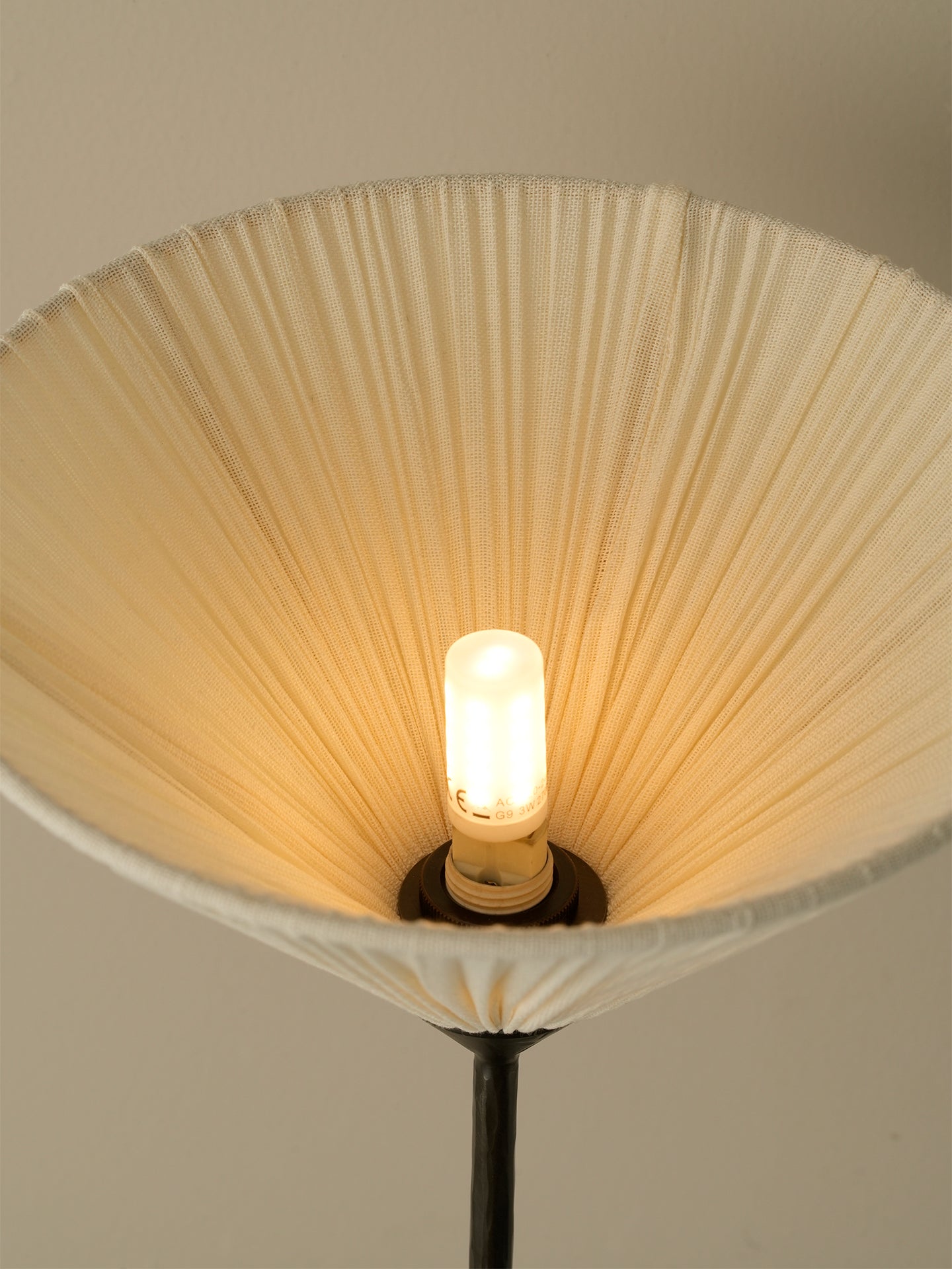 A close-up of a pleated fabric lampshade in a warm cream color, glowing with the light of an exposed filament bulb inside.