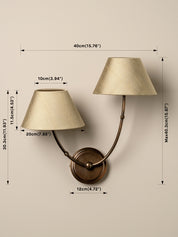 A wall-mounted two-light sconce with cream fabric shades and a bronze metal base, shown with overlaid dimension lines and measurements for width, height, and spacing on a beige wall.