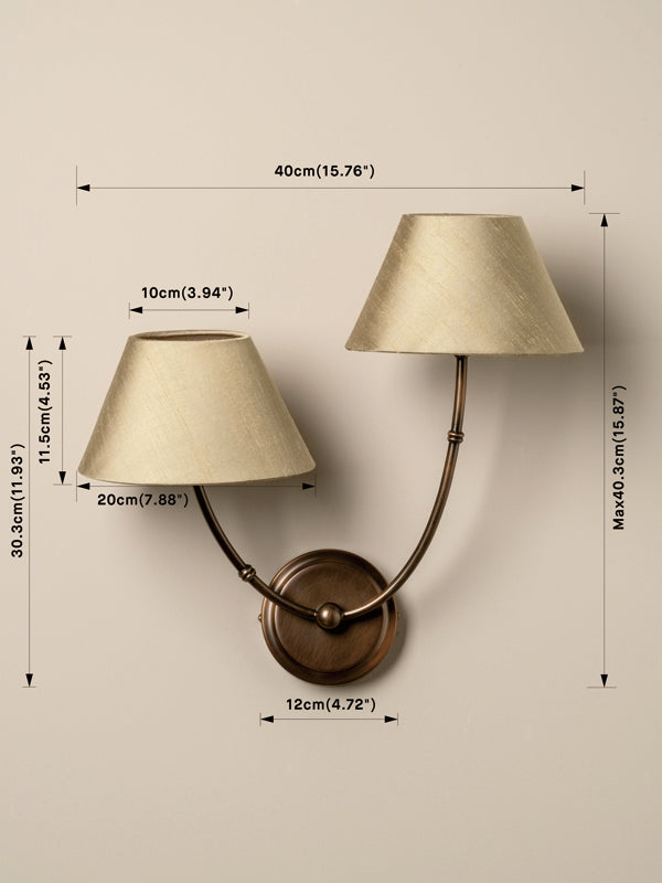 A wall-mounted two-light sconce with cream fabric shades and a bronze metal base, shown with overlaid dimension lines and measurements for width, height, and spacing on a beige wall.