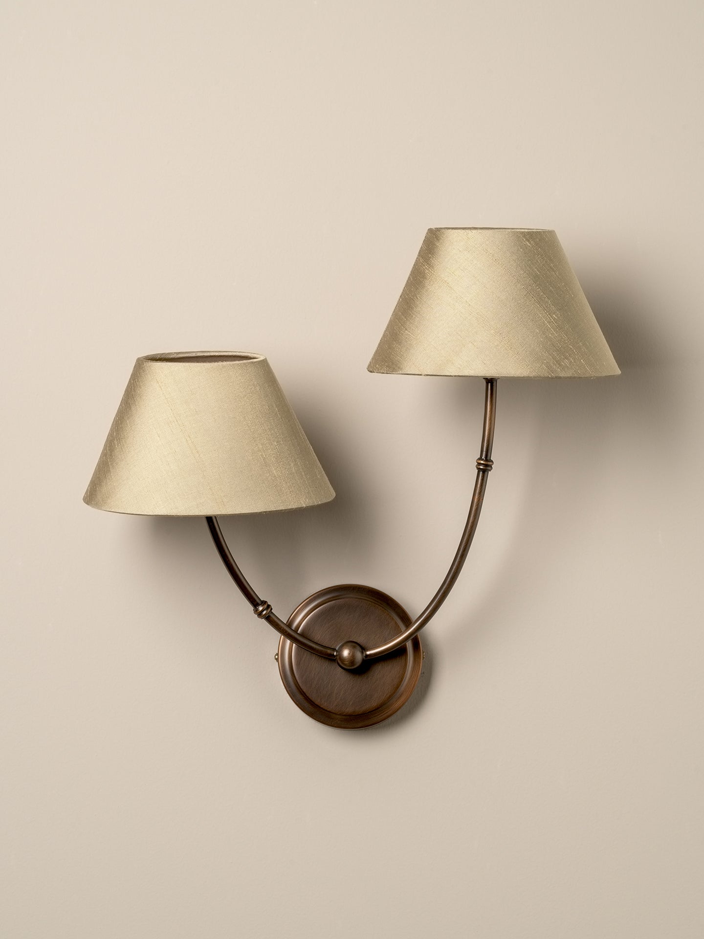 A double-arm wall sconce with tapered beige fabric shades and a round bronze wall mount, shown against a plain beige wall.