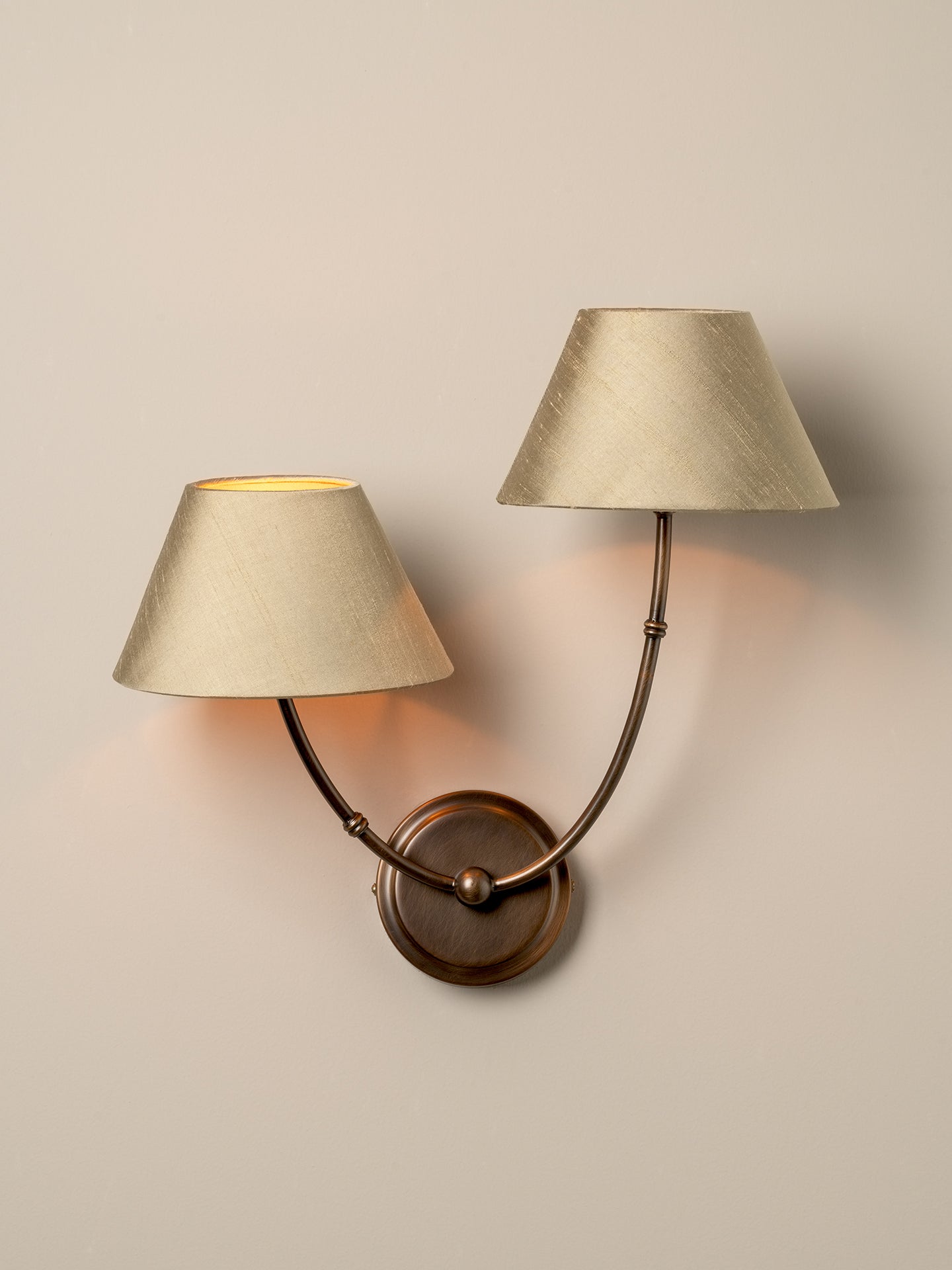 A double-arm wall sconce with textured beige fabric shades and a bronze-finished metal base, mounted on a warm beige wall and casting a soft glow.