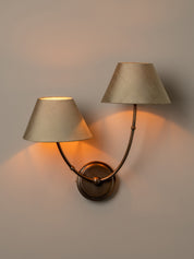 A two-light wall sconce with tapered beige fabric shades and curved bronze metal arms, mounted on a plain wall and illuminated with a warm glow.