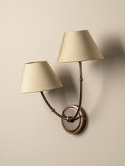 A double-arm wall sconce with tapered beige fabric shades and a bronze-finished metal frame, mounted on a plain beige wall.