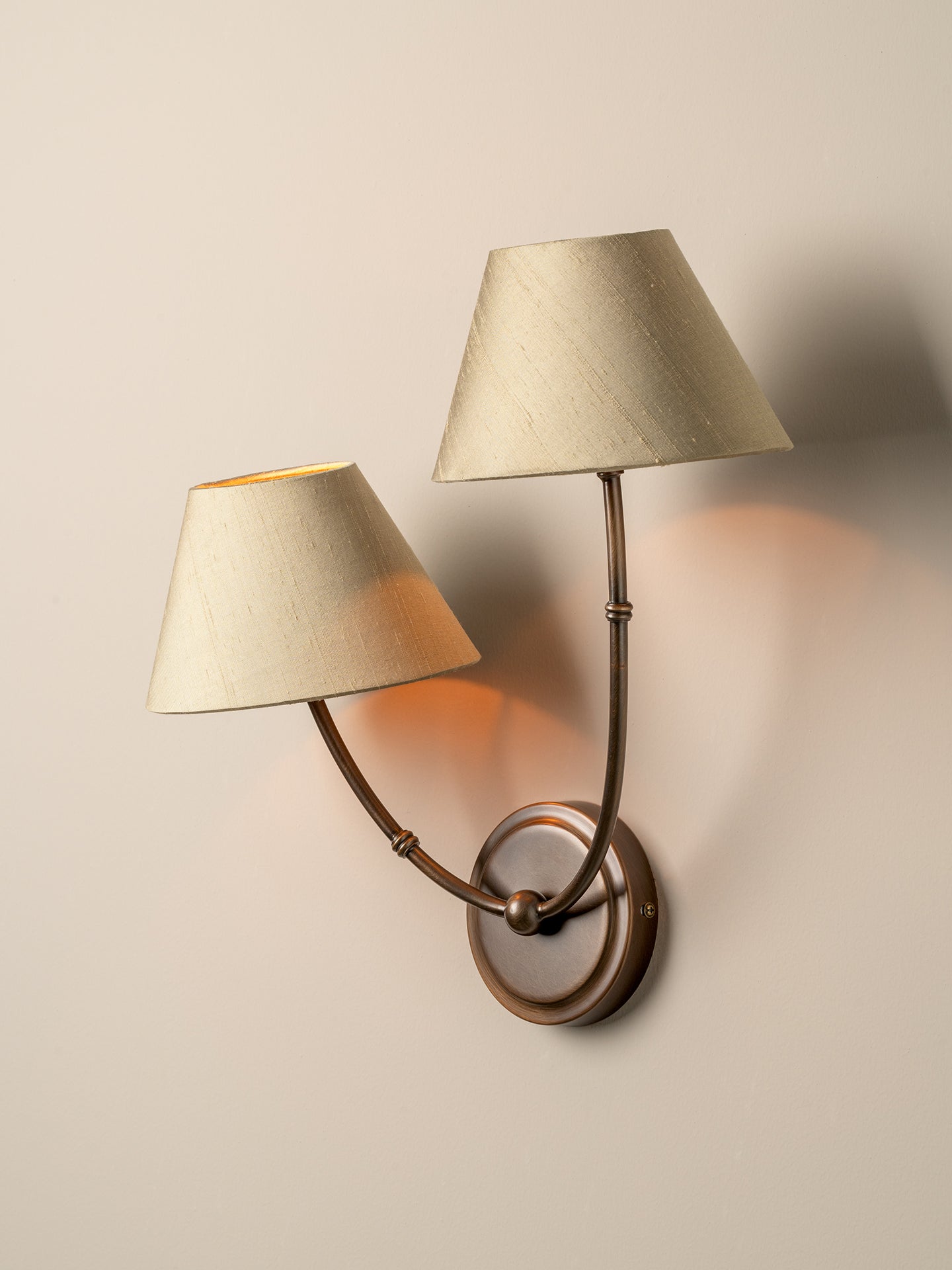 A two-light wall sconce with tapered beige fabric shades and a bronze-finished metal frame, mounted on a plain beige wall and casting warm light.