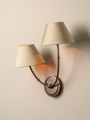 A two-light wall sconce with tapered beige fabric shades and a bronze-finished metal frame, mounted on a plain beige wall and casting warm light.