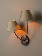 A two-light wall sconce with tapered beige fabric shades and a dark metal frame, shown lit against a plain wall.