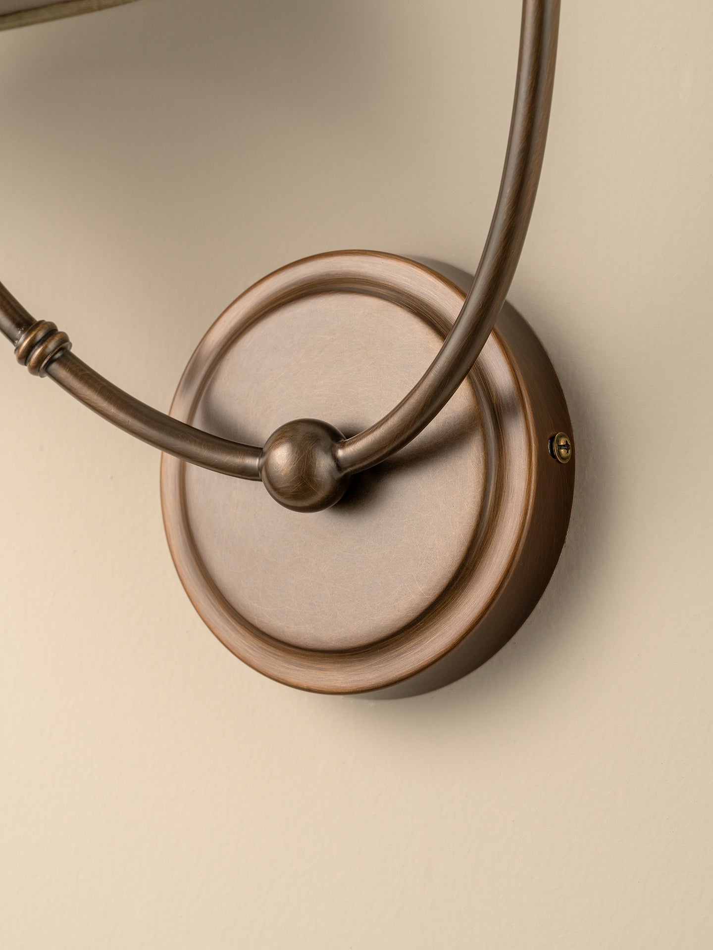 Close-up of a wall-mounted light fixture with a round bronze backplate and curved metal arms, shown against a warm beige wall.