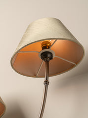 A close-up of a floor lamp shade with a warm beige fabric exterior and a metallic bronze interior, illuminated by an oncoming light bulb. The lamp has a dark metal central collar and a bronze-finished pole extending downward.