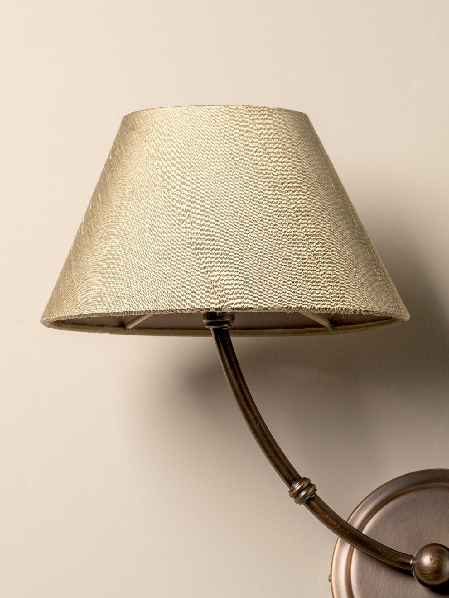 A close-up of a wall-mounted sconce with a tapered beige fabric shade and a dark curved metal arm, shown against a plain beige wall.