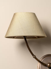 A close-up of a wall-mounted sconce with a tapered beige fabric shade and a dark curved metal arm, shown against a plain beige wall.