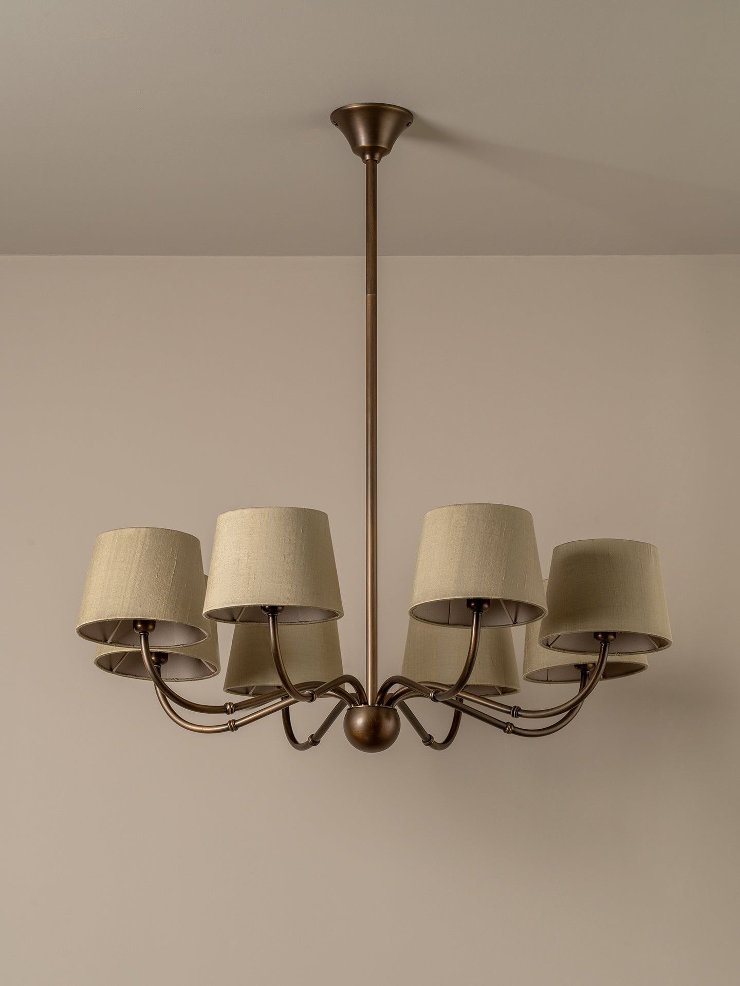 A six-light chandelier with a dark bronze metal frame and beige fabric drum shades, shown hanging against a neutral beige wall and ceiling.