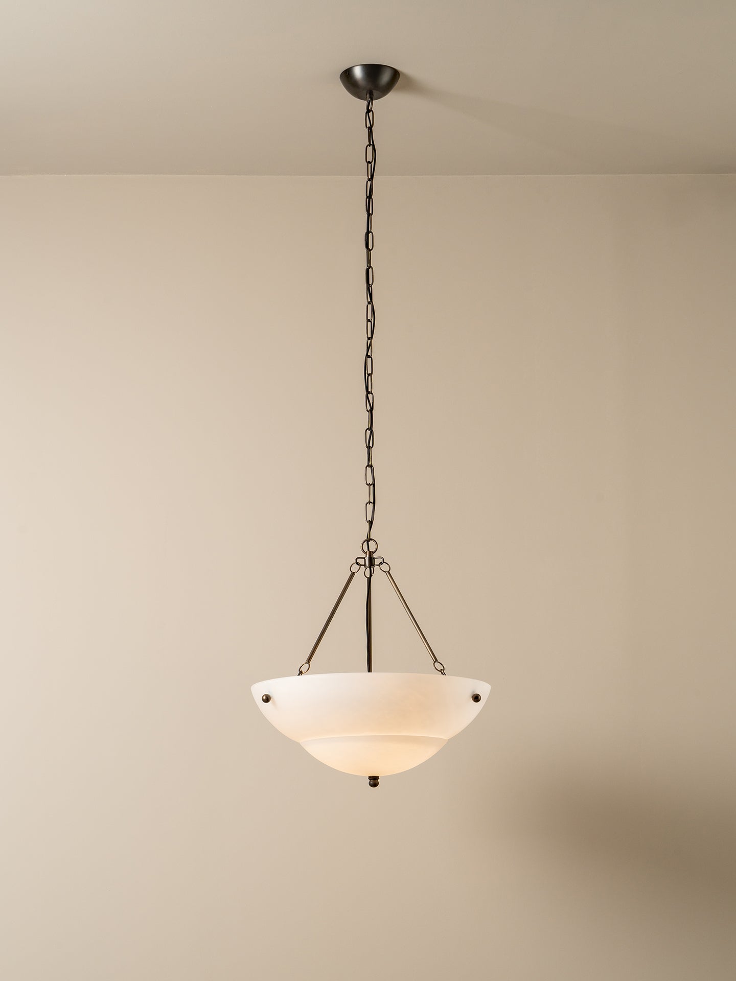 A modern pendant light with a frosted white glass bowl shade and a dark metal chain and canopy, hanging against a plain beige wall.
