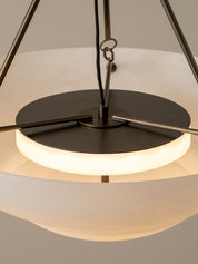 A close-up of a modern pendant light with a glowing warm white frosted glass globe shade, a matte black circular top plate, and brushed metal suspension rods against a neutral beige background.