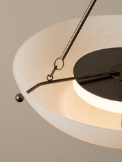 Close-up detail of a modern pendant light with a frosted white circular shade, a black circular ceiling mount, and brushed brass metal rods and hardware against a warm neutral background.