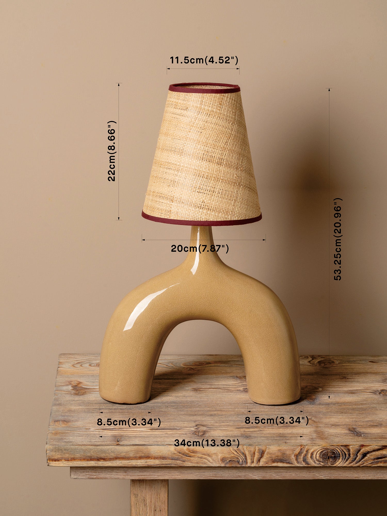 A beige ceramic table lamp with a curved base and a textured beige shade trimmed in dark red, shown on a rustic wooden table against a beige wall with overlaid dimension labels.