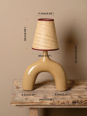 A beige ceramic table lamp with a curved base and a textured beige shade trimmed in dark red, shown on a rustic wooden table against a beige wall with overlaid dimension labels.