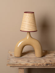 A sculptural table lamp with a glossy beige ceramic arch-shaped base and a textured beige woven lampshade trimmed in dark red, displayed on a rustic wooden table against a solid beige background.