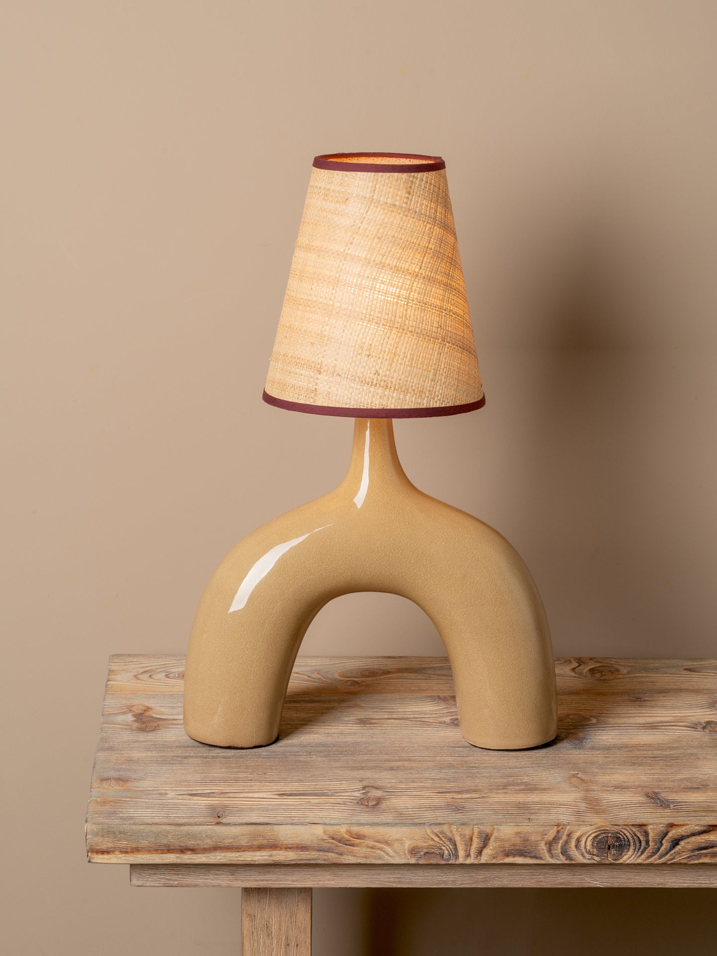 A sculptural table lamp with a glossy beige ceramic arch base and a tapered woven beige shade trimmed in dark red-brown, displayed on a rustic wooden table against a warm beige wall.