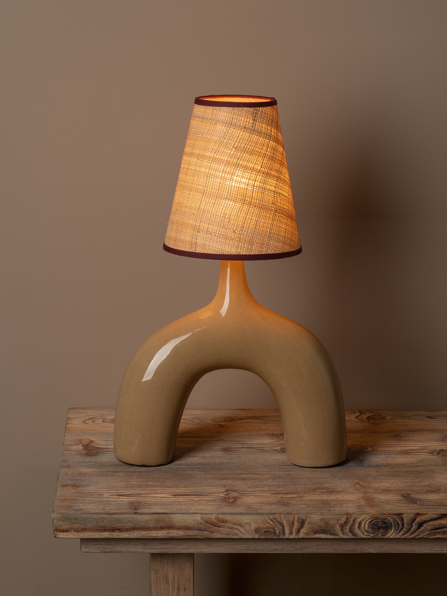 A sculptural table lamp with a glossy beige ceramic base and a textured beige fabric shade, illuminated with a warm glow, set on a rustic wooden table against a solid taupe background.