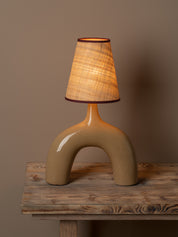 A sculptural table lamp with a glossy beige ceramic base and a textured beige fabric shade, illuminated with a warm glow, set on a rustic wooden table against a solid taupe background.