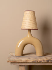 A sculptural table lamp with a glossy beige ceramic arch-shaped base and a textured beige woven shade, accented with dark red trim, displayed on a rustic wooden table against a solid beige background.