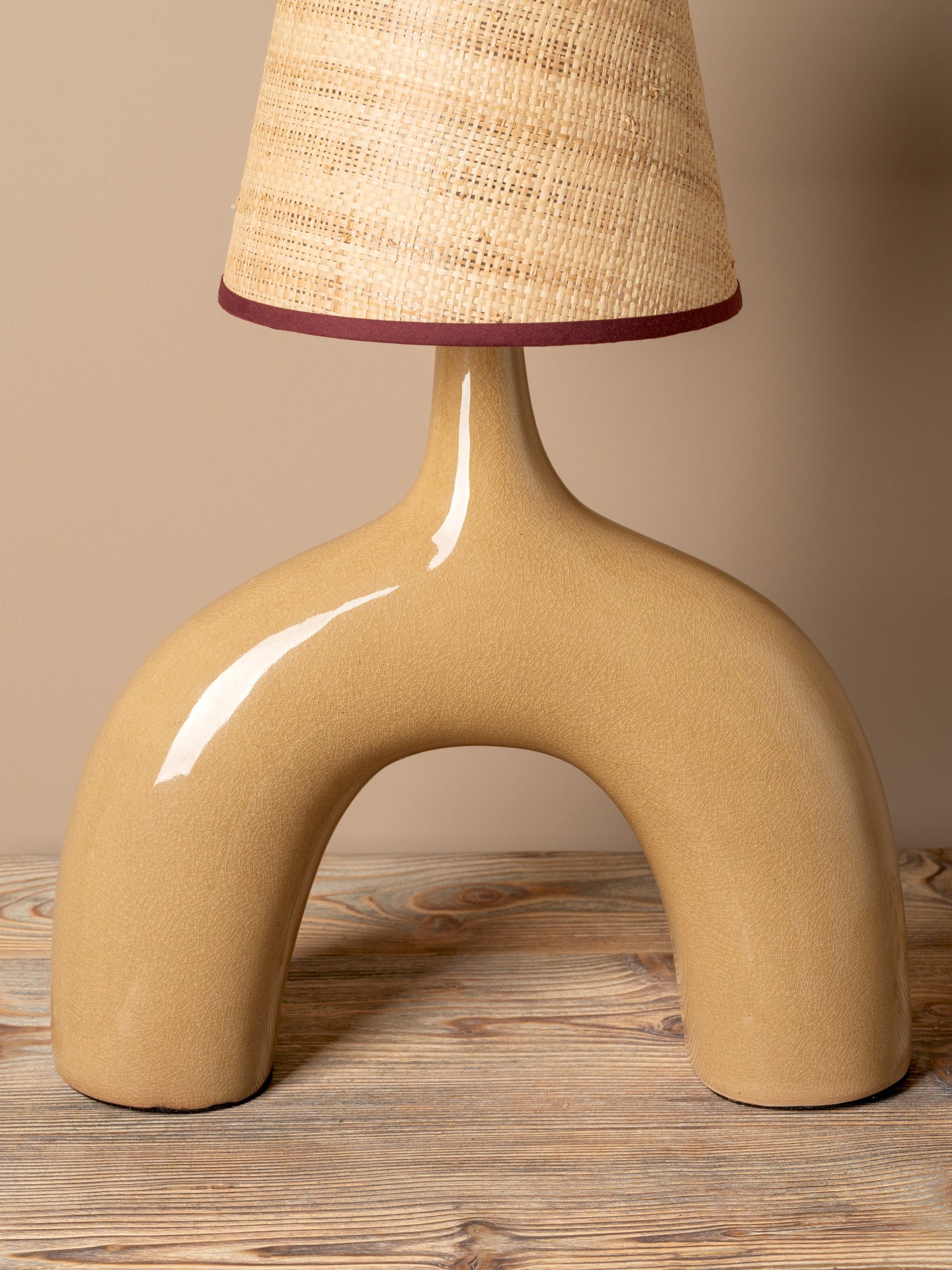 A modern table lamp with a glossy, curved beige ceramic base and a textured beige woven lampshade accented by a dark burgundy trim, set on a rustic wooden surface against a solid beige background.