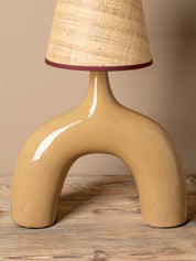 A modern table lamp with a glossy, curved beige ceramic base and a textured beige woven lampshade accented by a dark burgundy trim, set on a rustic wooden surface against a solid beige background.
